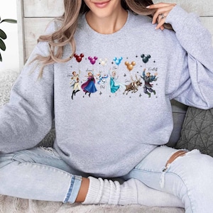 May include: A light gray sweatshirt featuring a colorful graphic of animated characters holding Mickey Mouse-shaped balloons. The characters include Hans, Anna, Olaf, Elsa, Sven, and Kristoff. The names of the characters are written above them.