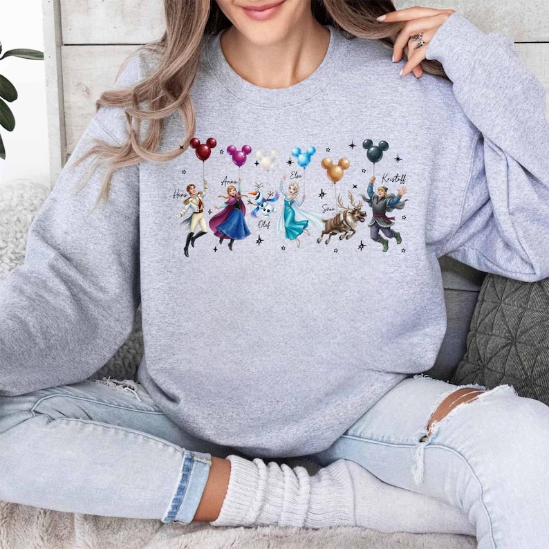May include: A light gray sweatshirt featuring a colorful graphic of animated characters holding Mickey Mouse-shaped balloons. The characters include Hans, Anna, Olaf, Elsa, Sven, and Kristoff. The names of the characters are written above them.