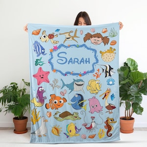 Può includere: Una coperta in pile azzurro con un motivo ripetuto di creature marine in stile cartone animato, tra cui uno squalo, un pesce pagliaccio, una tartaruga marina e una stella marina, con il nome Sarah scritto in blu scuro al centro.