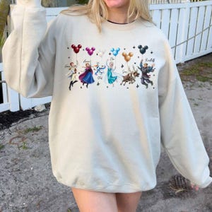 May include: Beige sweatshirt featuring a colorful graphic of characters from the movie Frozen, each holding a balloon. The characters include Anna, Elsa, Olaf, Sven, and Kristoff. The balloons are in various colors, including red, purple, blue, and black.