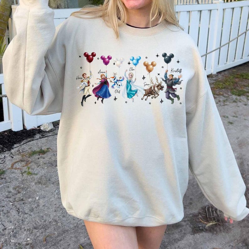 May include: Beige sweatshirt featuring a colorful graphic of characters from the movie Frozen, each holding a balloon. The characters include Anna, Elsa, Olaf, Sven, and Kristoff. The balloons are in various colors, including red, purple, blue, and black.