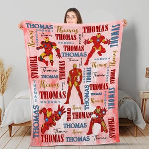 May include: A red and gold blanket featuring multiple illustrations of a superhero in action poses. The blanket has the name "Thomas" printed in various fonts and colors across a pink watercolor background.