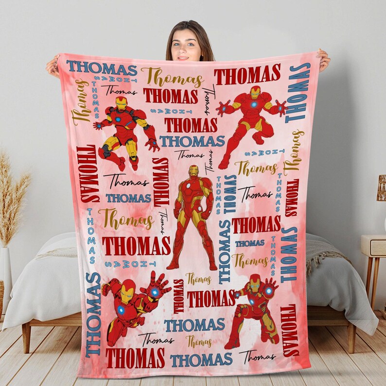 May include: A red and gold blanket featuring multiple illustrations of a superhero in action poses. The blanket has the name "Thomas" printed in various fonts and colors across a pink watercolor background.