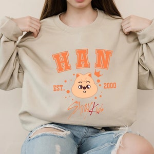May include: A beige sweatshirt featuring orange text and graphics. The word "HAN" is printed across the chest in a collegiate font. Below is a cartoon character with a crown, the text "EST. 2000", and the words "Stray Kids".