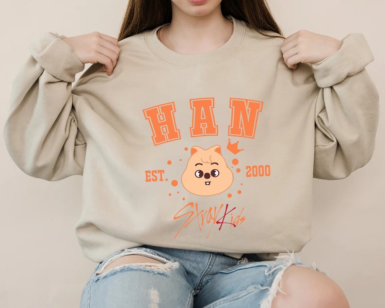 May include: A beige sweatshirt featuring orange text and graphics. The word "HAN" is printed across the chest in a collegiate font. Below is a cartoon character with a crown, the text "EST. 2000", and the words "Stray Kids".
