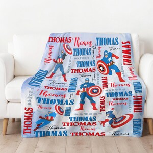 May include: A blue and white blanket featuring illustrations of a superhero with a shield. The blanket has the name "Thomas" printed in red and white, along with the superhero's image. The blanket is draped over a white couch.