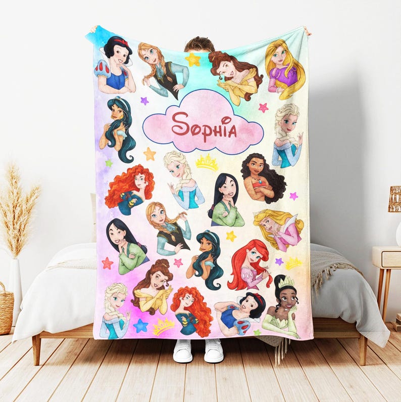 May include: A personalized blanket featuring cartoon illustrations of various princesses. The blanket is predominantly white with a colorful design, including stars and a cloud with the name "Sophia" in pink script. The princesses are depicted in their signature outfits.