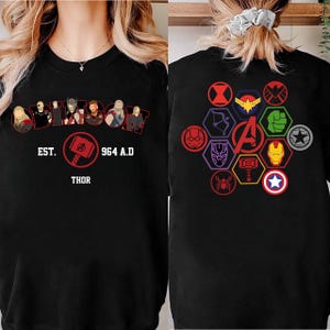Puede incluir: Sudadera negra con un dise&ntilde;o que presenta la palabra "Thor", el texto "EST. 964 A.D." e ilustraciones de personajes. La parte trasera de la sudadera muestra varios s&iacute;mbolos de superh&eacute;roes y la letra "A" en el centro.