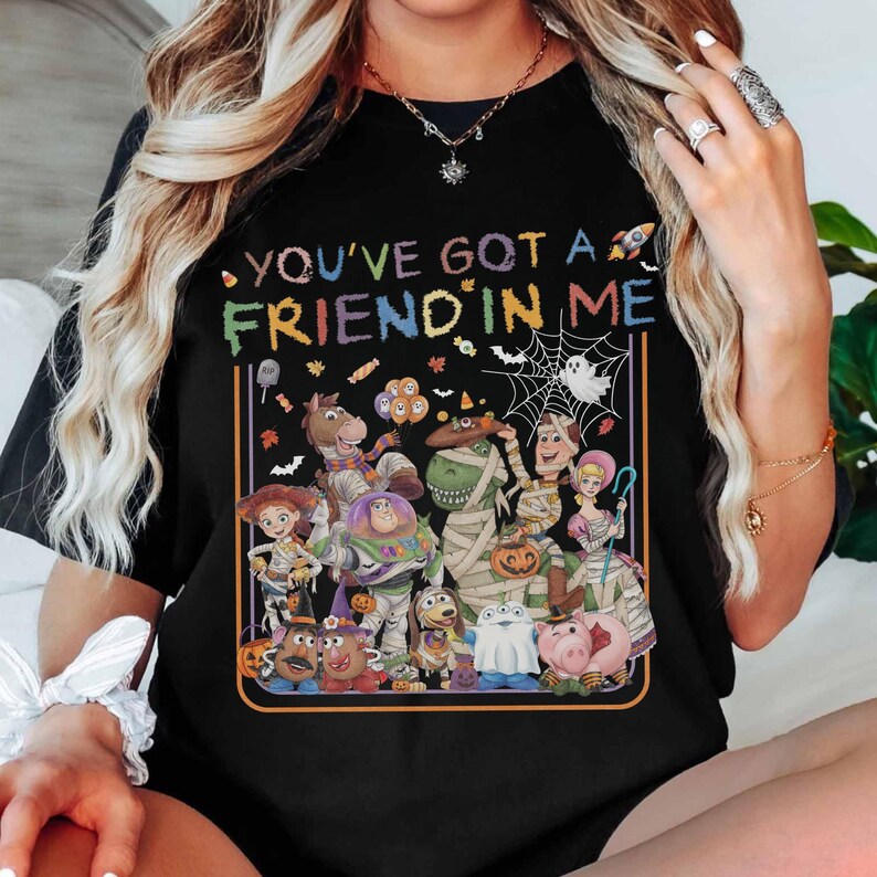 K&ouml;nnte beinhalten: Ein schwarzes T-Shirt mit dem Text "YOU'VE GOT A FRIEND IN ME" in bunten Buchstaben &uuml;ber einer grafischen Illustration verschiedener Zeichentrickfiguren in Halloween-Kost&uuml;men, darunter eine Mumie, ein Geist und ein K&uuml;rbis.