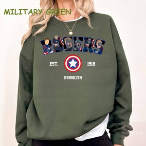 May include: Military green sweatshirt with the word "AVENGERS" in a stylized font, featuring character illustrations. Below, a Captain America shield graphic is centered above the text "EST. 1918 BROOKLYN". The sweatshirt is a solid colour.
