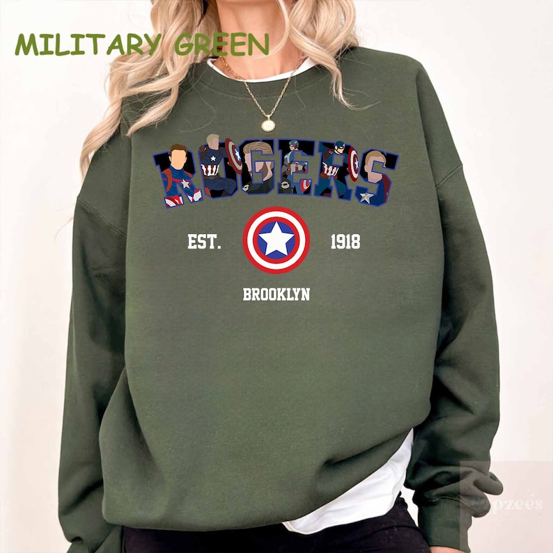 May include: Military green sweatshirt with the word "AVENGERS" in a stylized font, featuring character illustrations. Below, a Captain America shield graphic is centered above the text "EST. 1918 BROOKLYN". The sweatshirt is a solid colour.