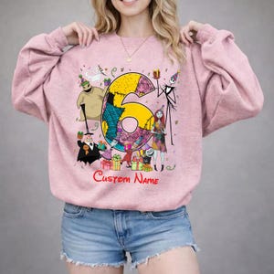 May include: A light pink crewneck sweatshirt featuring a large yellow number six graphic surrounded by characters from The Nightmare Before Christmas, including Jack Skellington, Sally, and Oogie Boogie, with the red text Custom Name below.