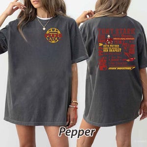 May include: Dark gray t-shirt with a graphic on the front and back. The front features a yellow and red design. The back has red text that reads "Tony Stark" and "Stark Industries". The shirt is a comfortable, casual style.