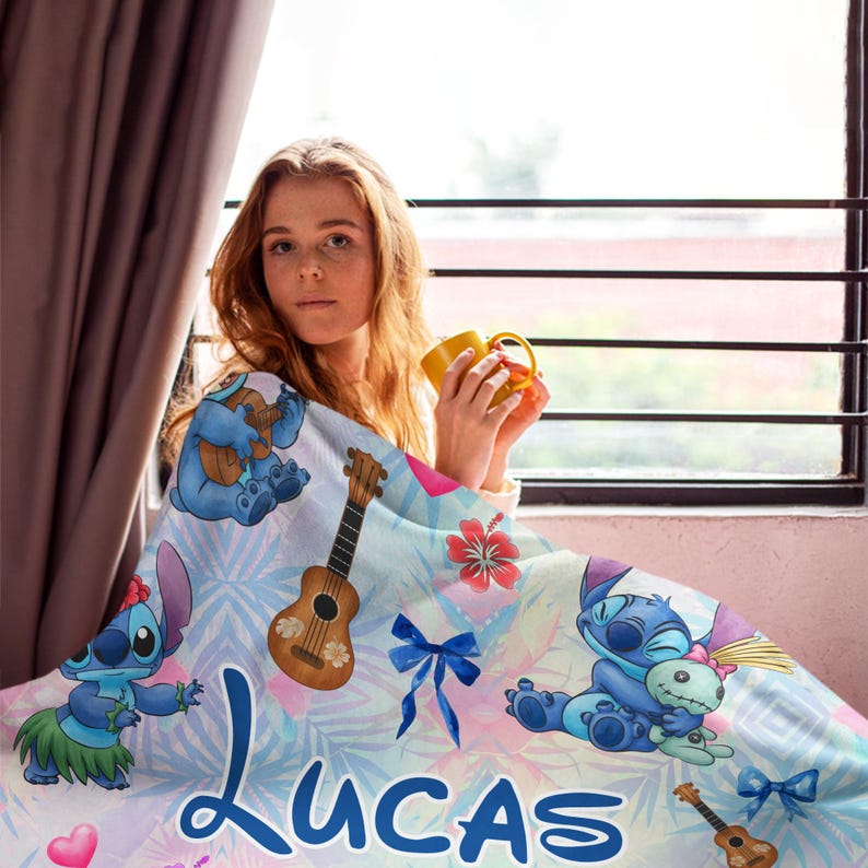 Op de afbeelding: Een lichtblauwe deken met stripfiguren, ukeleles en tropische bloemen. De deken heeft de naam "Lucas" erop gedrukt. Een persoon is in de deken gewikkeld en houdt een gele mok vast.