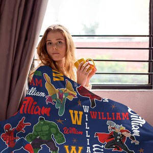 May include: A navy blue blanket featuring illustrations of Marvel superheroes, including Spider-Man, Hulk, and Thor. The blanket also displays the name "William" in various fonts and colors. The blanket is draped over a person.