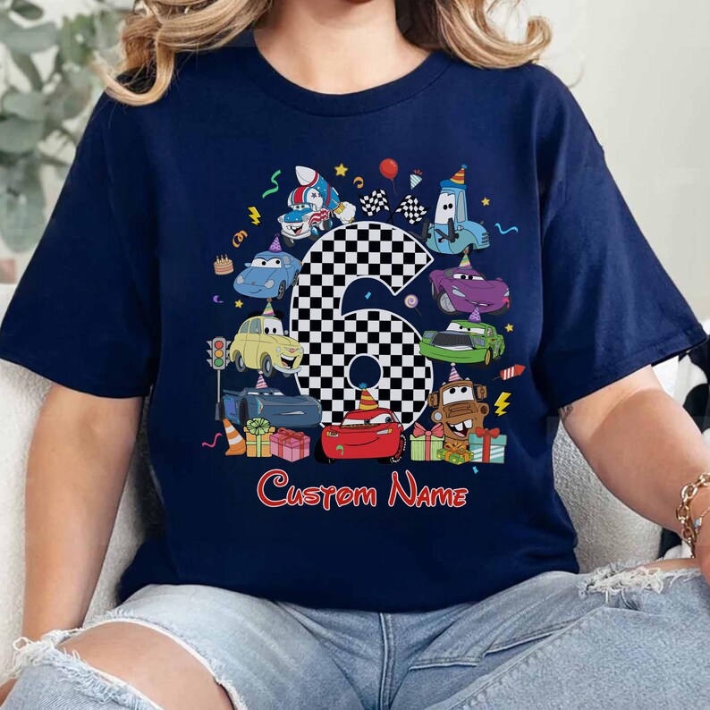 May include: Navy blue t-shirt with a vibrant cartoon design of race cars, a checkered number six, and the text "Custom Name". The design incorporates birthday elements such as confetti, balloons, and gifts. A fun, celebratory theme.
