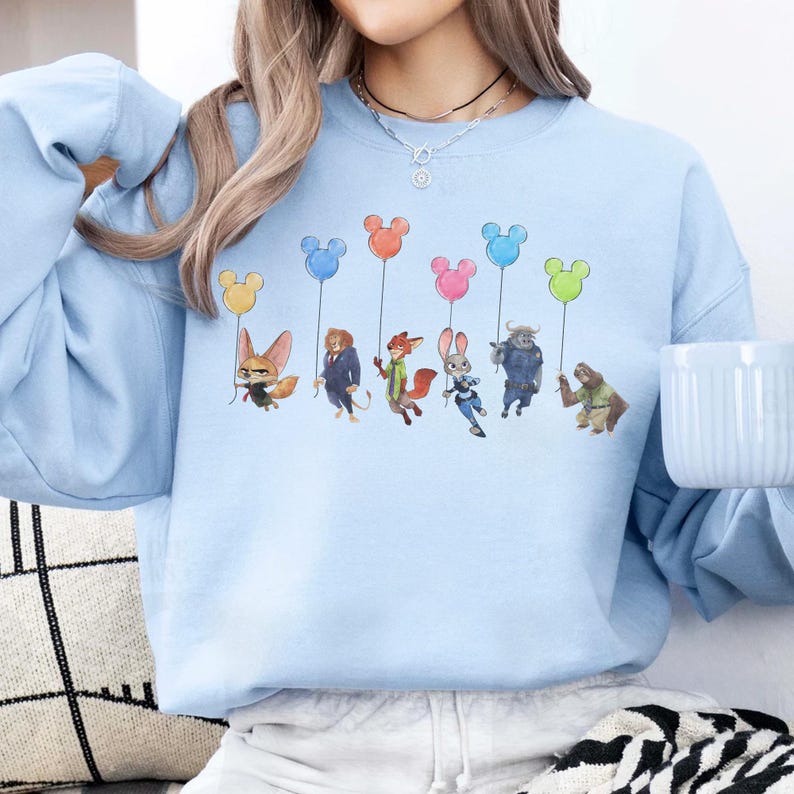 May include: Light blue sweatshirt featuring a colorful graphic of cartoon characters holding Mickey Mouse-shaped balloons. The characters are from the animated movie Zootopia. The sweatshirt is a comfortable, casual style.