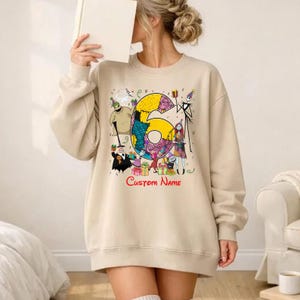 May include: A beige crewneck sweatshirt featuring a large graphic of the number six decorated with patterns, surrounded by various animated characters from The Nightmare Before Christmas, colorful gift boxes, and the red text Custom Name.
