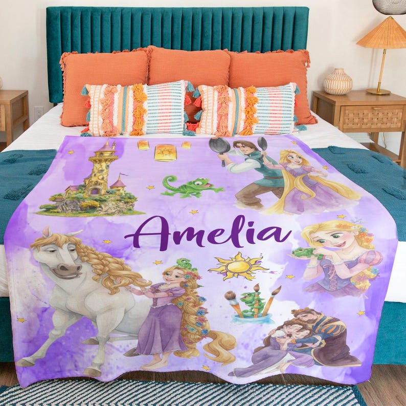 May include: A purple fleece blanket with fairytale princess illustrations, a horse, and a castle. The blanket has the name "Amelia" in a cursive font. The design includes a sun, frogs, and other characters.