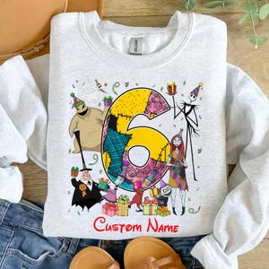 May include: A light gray crewneck sweatshirt featuring a large yellow number six surrounded by various animated characters from The Nightmare Before Christmas, including Jack Skellington, Sally, and Oogie Boogie, with the text Custom Name below.