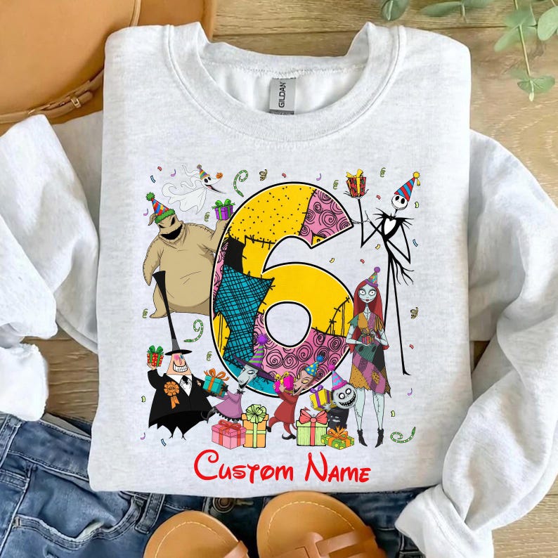 May include: A light gray crewneck sweatshirt featuring a large yellow number six surrounded by various animated characters from The Nightmare Before Christmas, including Jack Skellington, Sally, and Oogie Boogie, with the text Custom Name below.