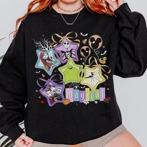 May include: Black crew neck sweatshirt with a vibrant graphic. The design incorporates star-shaped characters, balloons, and the word "magical" in a playful font. The theme is a Halloween or fantasy design.