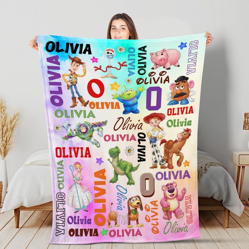 Op de afbeelding: Een rechthoekige fleece deken met een kleurrijke print van diverse geanimeerde speelgoedfiguren, waaronder een cowboy, space ranger, cowgirl, dinosaurus en een varken. De naam OLIVIA is herhaaldelijk in verschillende lettertypes en kleuren gedrukt.