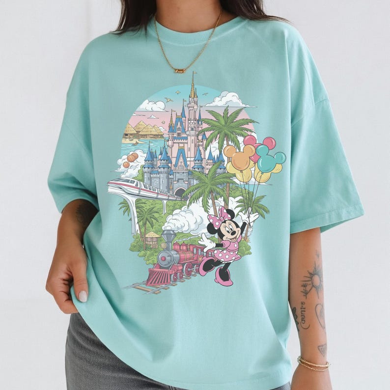 以下が含まれることがあります： 水色のTシャツ。お城、電車、ヤシの木、ミニーマウスが風船を持っているカラフルなグラフィックが特徴です。デザインは円形のフレーム内に配置されています。このシャツは半袖で、ゆったりとしたフィット感です。