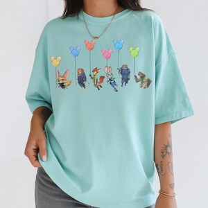 May include: A light blue t-shirt featuring a graphic of characters from the animated film "Zootopia" holding Mickey Mouse-shaped balloons in various colors. The shirt has a relaxed fit and short sleeves.