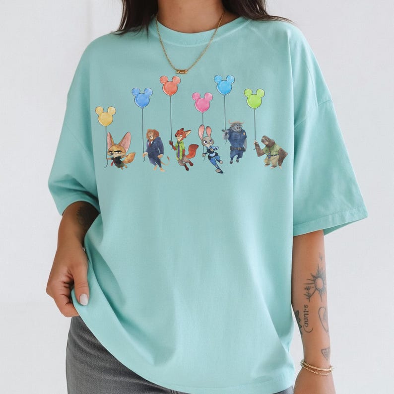May include: A light blue t-shirt featuring a graphic of characters from the animated film "Zootopia" holding Mickey Mouse-shaped balloons in various colors. The shirt has a relaxed fit and short sleeves.