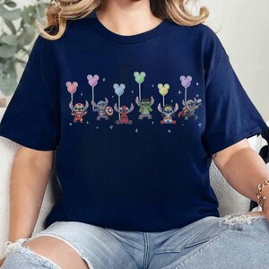 May include: Navy blue t-shirt featuring a row of cartoon characters dressed as superheroes, each holding a Mickey Mouse-shaped balloon. The characters are in various colors and poses, with small star accents. The shirt is a casual wear item.