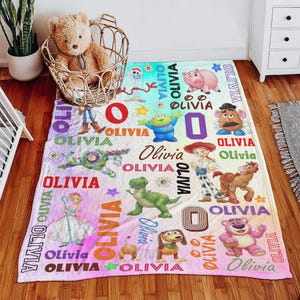 Op de afbeelding: Een rechthoekige fleece deken met een herhalend patroon van diverse tekenfilmfiguren, waaronder een groene dinosaurus, een space ranger, een cowgirl, een varken en een aardappelfiguur, afgewisseld met de naam Olivia in diverse kleuren.