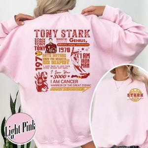 May include: Light pink sweatshirt featuring a graphic of Tony Stark with text including "Genius, billionaire, philanthropist" and "I am Iron Man." The design incorporates red and gold elements, with the Stark Industries logo on the front.
