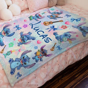 Op de afbeelding: Een gepersonaliseerde blauw-witte deken met het personage Stitch uit Lilo & Stitch. De deken heeft meerdere afbeeldingen van Stitch, ukeleles en tropische bloemen. De naam "Lucas" staat in grote blauwe letters op de deken.
