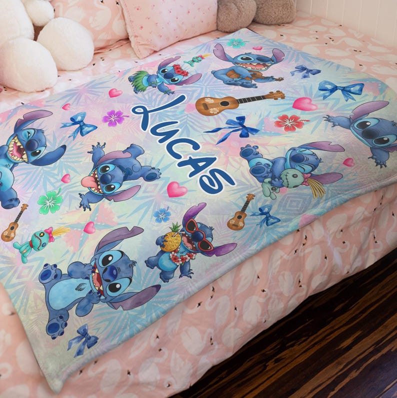 Op de afbeelding: Een gepersonaliseerde blauw-witte deken met het personage Stitch uit Lilo & Stitch. De deken heeft meerdere afbeeldingen van Stitch, ukeleles en tropische bloemen. De naam "Lucas" staat in grote blauwe letters op de deken.