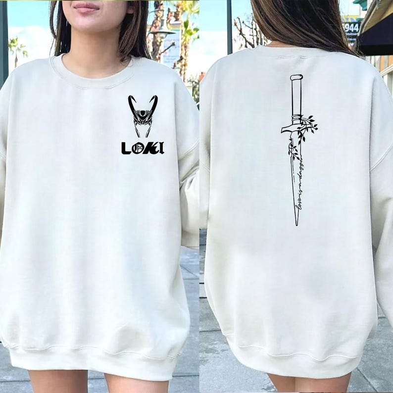 May include: Cream-colored sweatshirt featuring a black graphic of Loki's helmet and the name "LOKI" on the front. The back displays a black illustration of a dagger with floral accents and the text "Always a little mischief."