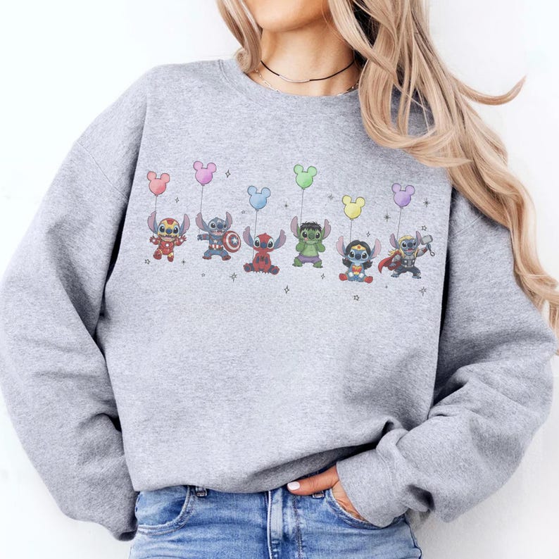 May include: A light gray sweatshirt featuring a colorful graphic of cartoon characters dressed as superheroes, each holding a Mickey Mouse-shaped balloon. The characters include Stitch, Captain America, Deadpool, Hulk, Wonder Woman, and Thor.