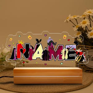 May include: A personalized night light with the name "NAME" in colorful letters, featuring Disney villains. The light sits on a wooden base and is connected to a power cord. The design includes hearts, crowns, and other decorative elements.