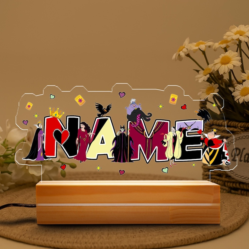 May include: A personalized night light with the name "NAME" in colorful letters, featuring Disney villains. The light sits on a wooden base and is connected to a power cord. The design includes hearts, crowns, and other decorative elements.