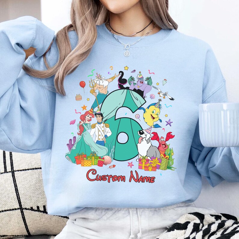 K&ouml;nnte beinhalten: Ein hellblaues Sweatshirt mit Rundhalsausschnitt und einer gro&szlig;en t&uuml;rkisfarbenen Zahl sechs, umgeben von Zeichentrickfiguren aus Arielle, die Meerjungfrau, darunter Arielle, Prinz Erik, Fabius, Sebastian, Triton und Ursula, mit dem Text Custom Name.