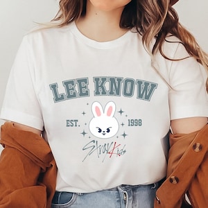 May include: White t-shirt featuring the text "LEE KNOW" in a blue-grey font, a cartoon bunny face, and the text "EST. 1998" and "Stray Kids". The shirt has a crew neck and short sleeves.