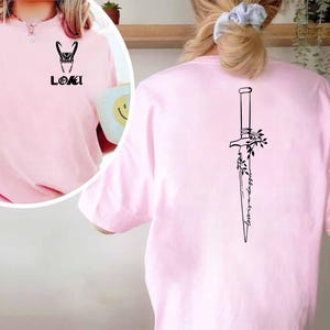 May include: A light pink t-shirt featuring a black graphic of Loki's helmet and the word "LOKI" on the front. The back of the shirt displays a black dagger design with the words "Always in trouble".