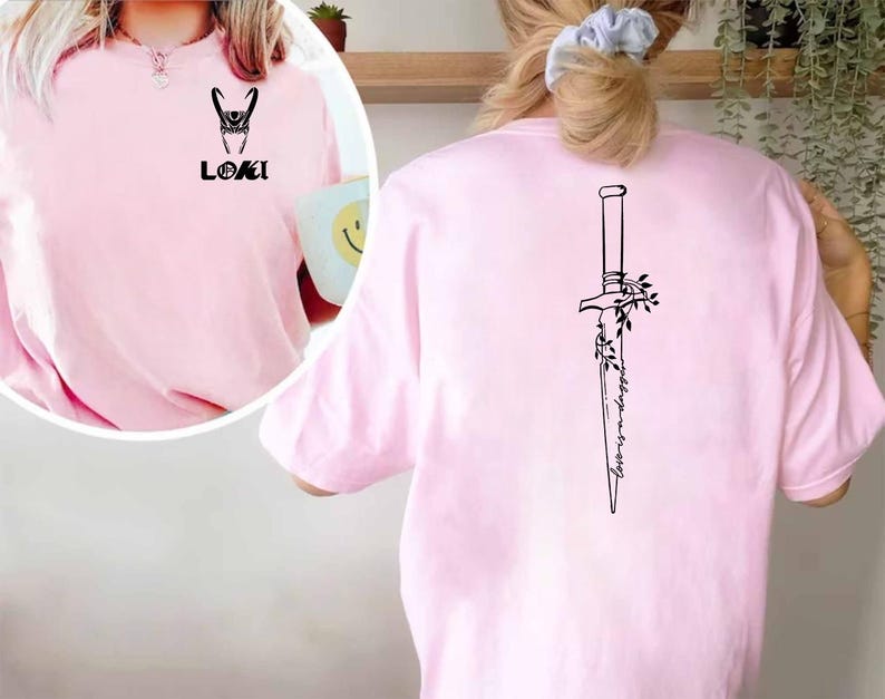 May include: A light pink t-shirt featuring a black graphic of Loki's helmet and the word "LOKI" on the front. The back of the shirt displays a black dagger design with the words "Always in trouble".