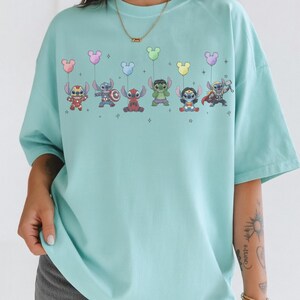 May include: A light blue t-shirt featuring a colorful graphic of cartoon characters holding balloon bouquets. The characters include Stitch, Hulk, and Thor. The shirt has a crew neck and short sleeves.