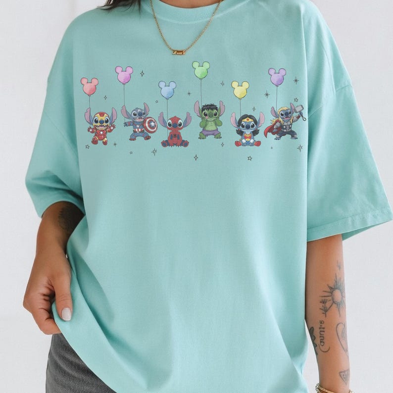 May include: A light blue t-shirt featuring a colorful graphic of cartoon characters holding balloon bouquets. The characters include Stitch, Hulk, and Thor. The shirt has a crew neck and short sleeves.