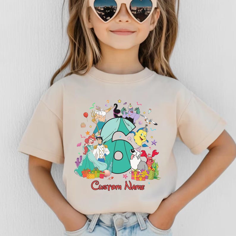 K&ouml;nnte beinhalten: Ein beiges Kurzarm-T-Shirt mit einer gro&szlig;en t&uuml;rkisfarbenen Zahl sechs, umgeben von bunten Zeichentrickfiguren aus Arielle, die Meerjungfrau, darunter Arielle, Prinz Erik, Fabius und Sebastian, mit dem roten Text Custom Name darunter gedruckt.
