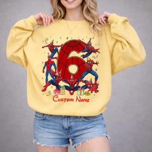 May include: A pale yellow crewneck sweatshirt featuring a large red number six with a spiderweb pattern. The design includes multiple Spider-Man figures wearing colourful party hats, surrounded by gift boxes and the text Custom Name.