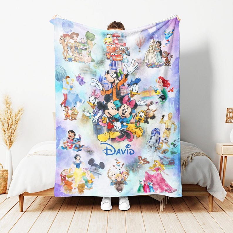 Op de afbeelding: Een kleurrijke deken met een collage van Disney-personages, waaronder Mickey en Minnie Mouse, Donald Duck en verschillende prinsessen. De deken heeft een aquarel-achtige achtergrond met de naam "David" in een scriptlettertype.