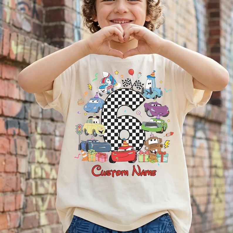 May include: A beige t-shirt with a colourful cartoon car design and the number 6 in a chequered pattern. The text "Custom Name" is written in red script below the cars. The shirt is for a child.