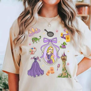 May include: Beige t-shirt featuring a collection of cartoon illustrations. The design includes a princess portrait, a tower, a dress, a frying pan, and other related elements in purple, yellow, and green.